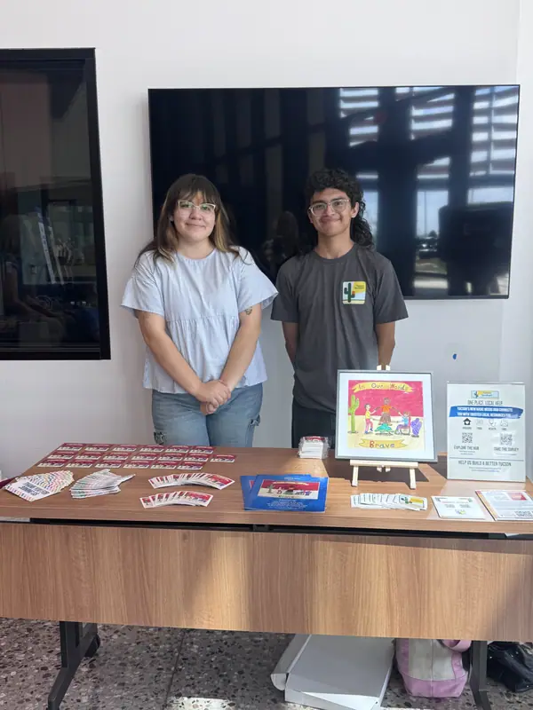 Two people stand behind a table of printed materials and artwork at the Tucson event.