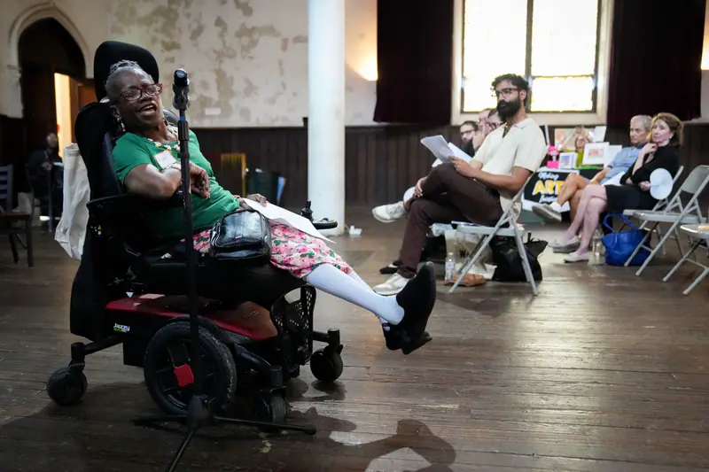 A storyteller in a power wheelchair speaks into a microphone while the audience listens.
