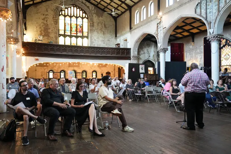 The audience seated in rows inside 2640 Space, a tall hall with arched windows.