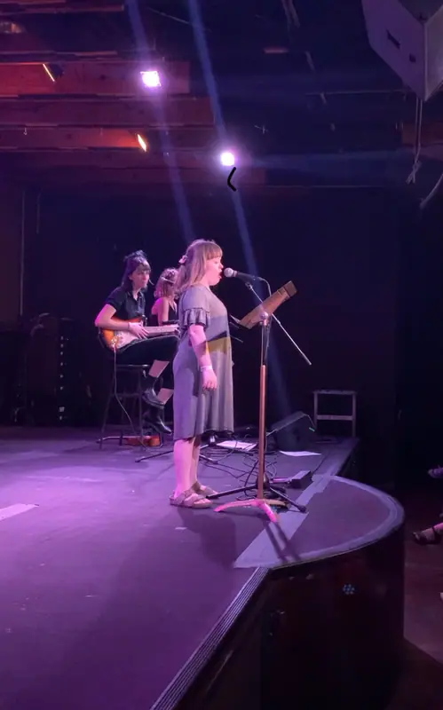 A storyteller stands at a microphone on stage under purple light, with musicians behind her.