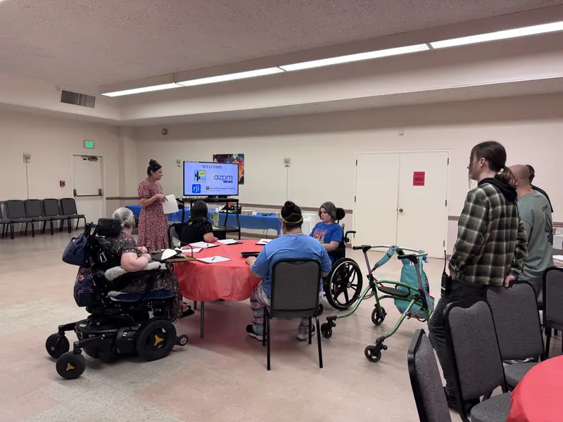 A workshop in progress: participants around tables, several using wheelchairs, facing a presenter and a screen.